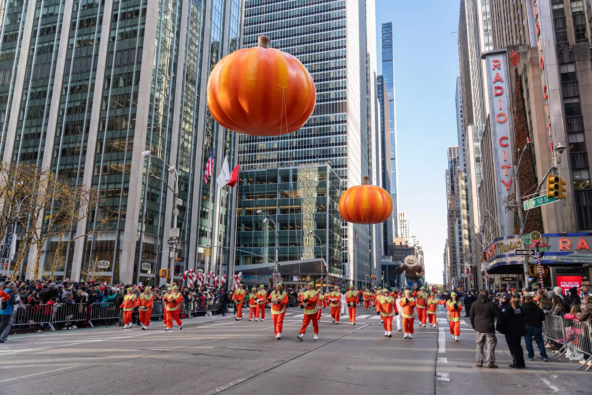 Massive balloon pumpkins float above a city street lined with high-rise buildings held onto by people in a parade dressed in pumpkin colors.