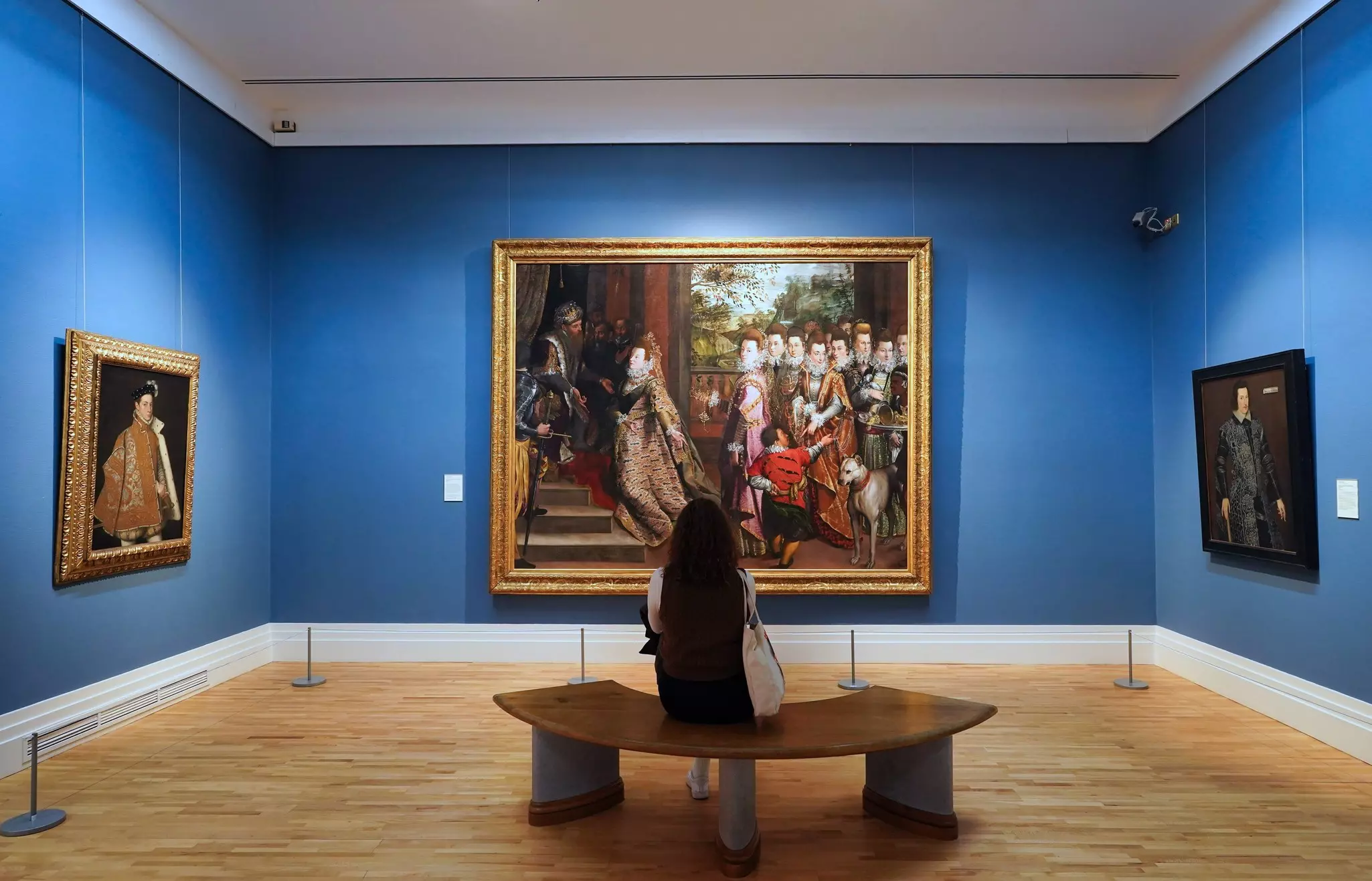 A woman sits on a bench and looks at a large painting in a museum gallery. The walls are painted blue.