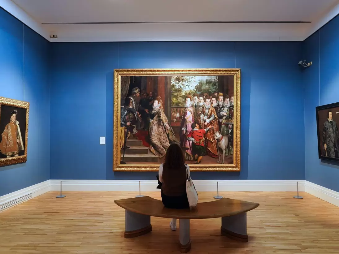 Interior of Ireland National Gallery of Art, Dublin, Ireland. Woman sitting in front of large painting against a blue gallery wall. 