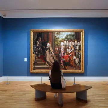 Interior of Ireland National Gallery of Art, Dublin, Ireland. Woman sitting in front of large painting against a blue gallery wall. 