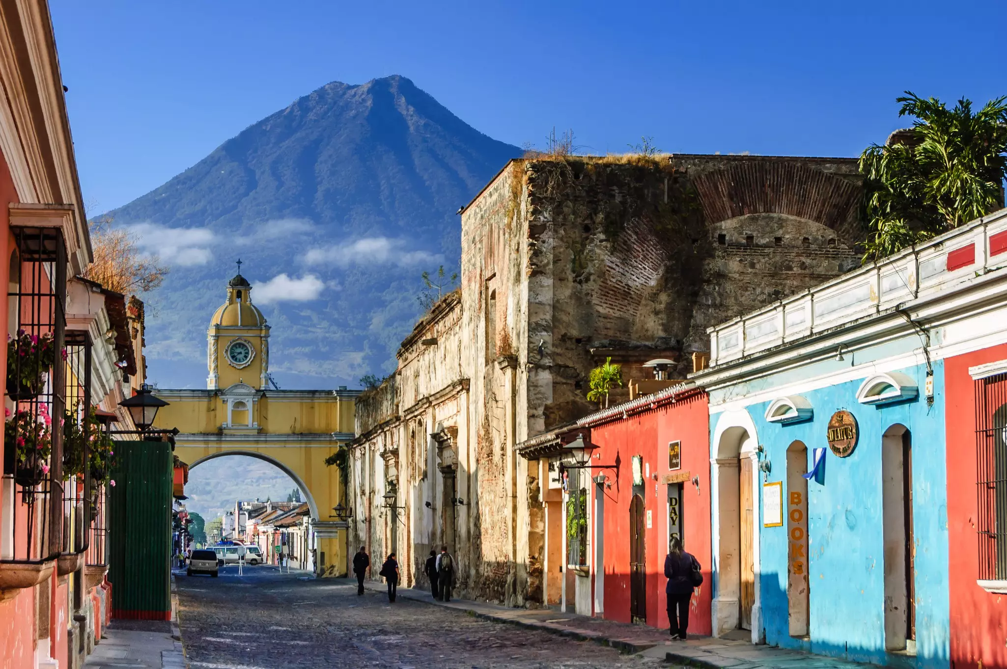 The pointed cone of a volcano rises above a low-rise city with pastel-colored buildings and an archway over a cobbled street.