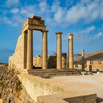 Standing sandstone columns and a stone stairway of an ancient temple on a cliff edge beneath a blue sky with white clouds.