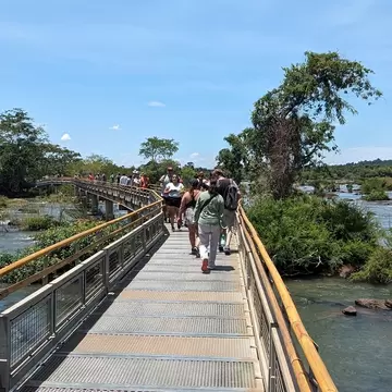 Visitors on a metal walkway with yellow handrails crossing rocky river channels and green islands under a blue sky