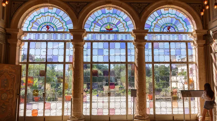 A woman looks at a plaque while standing in front of three arched floor to ceiling windows variously paned with colored stained glass