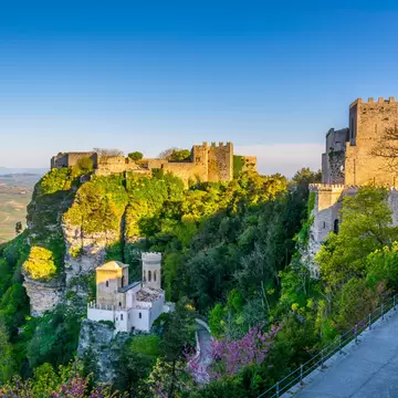 Medieval stone castle and towers on a green, wooded cliff in Erice, Sicily, lit by low sun, with farmland far below.