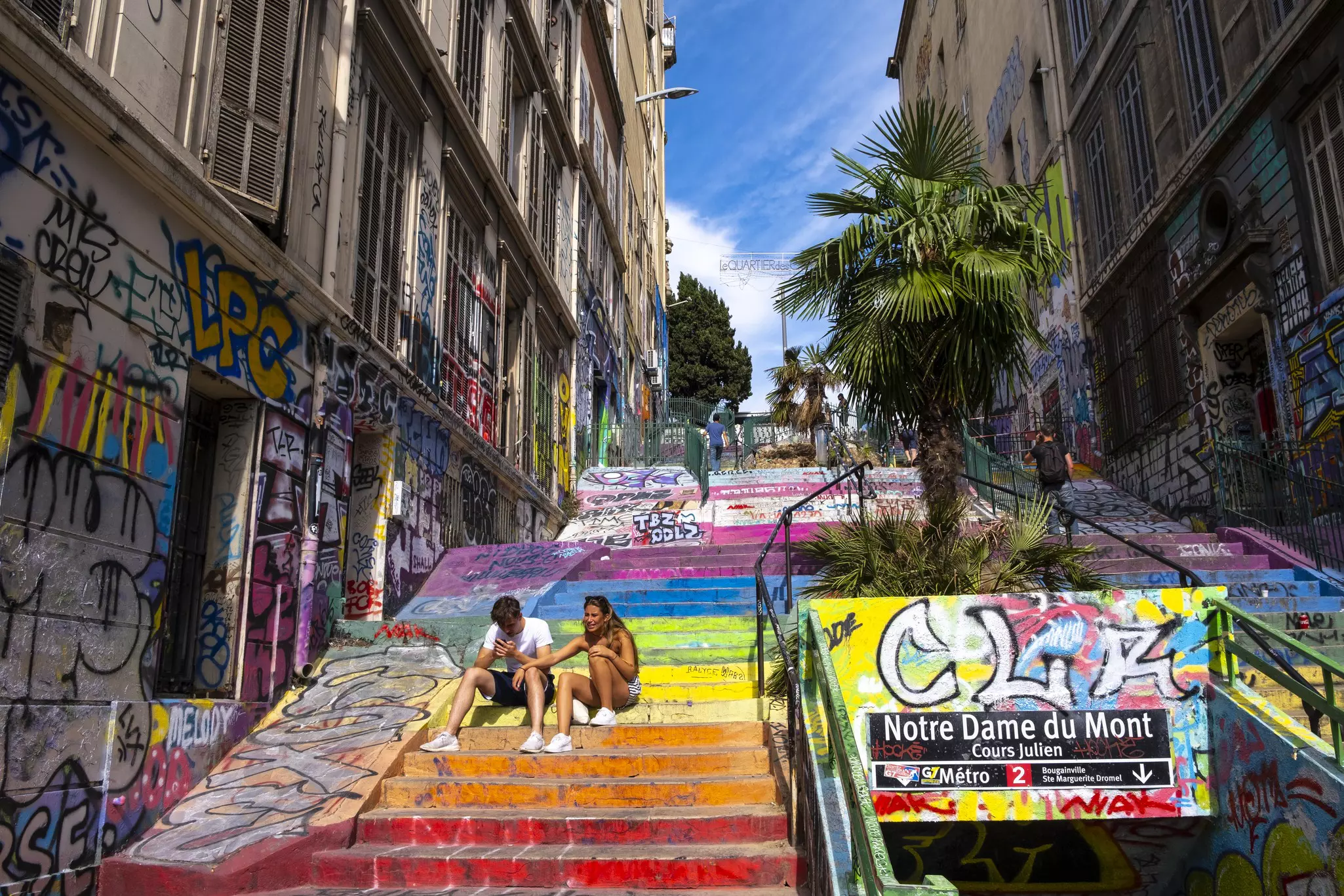 Two young people sit on an exterior staircase in a city with steps painted in the colors of the rainbow.