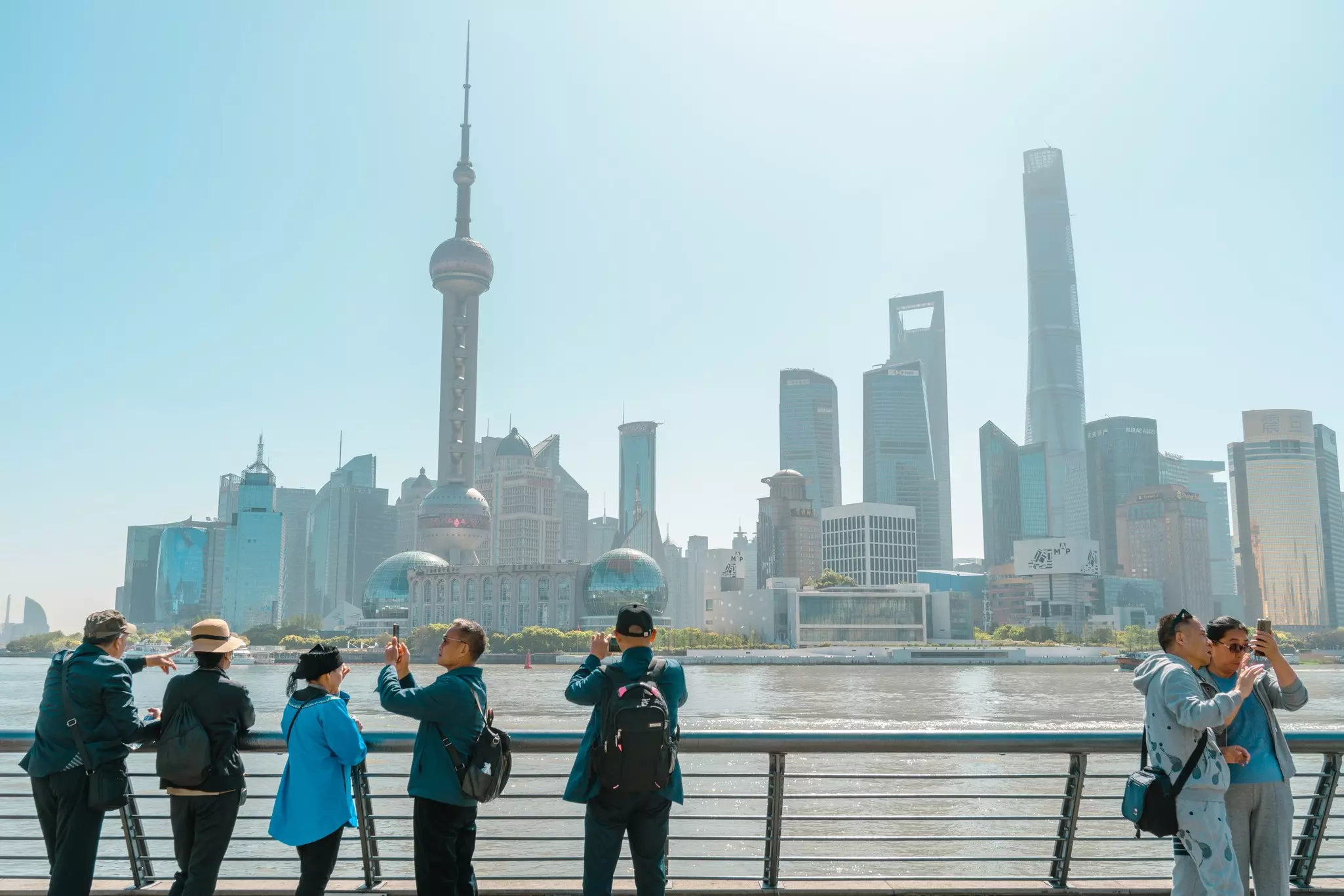 People stand near railings beside a river looking at the skyscrapers on the opposite side.