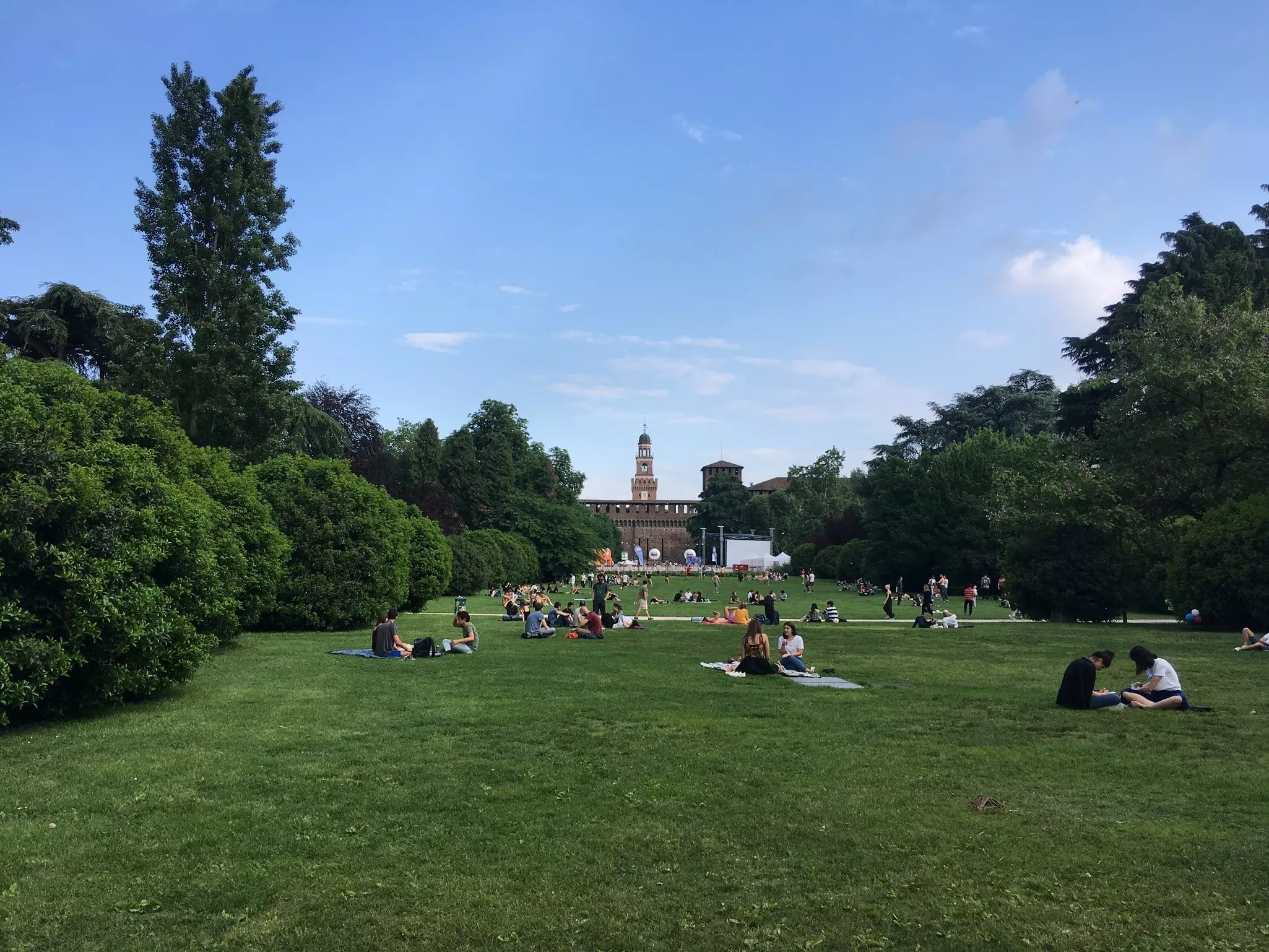 People in Milan sitting on a long lawn of green grass that extends to a building with a tower at the end.