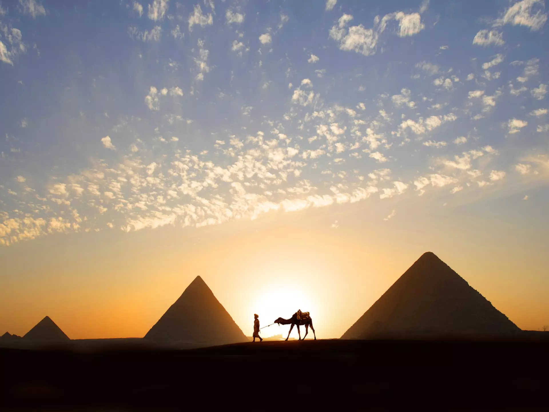 A single tour guide walking a camel at sunset by a huge stone pyramid in the desert