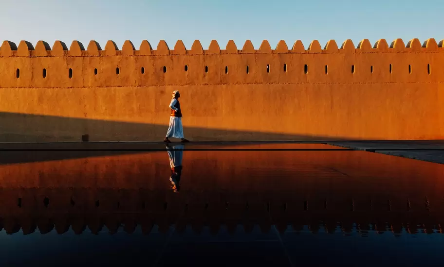 Man in a white robe walks beside a crenellated ochre fort wall in Abu Dhabi emirate, reflected in a dark pool