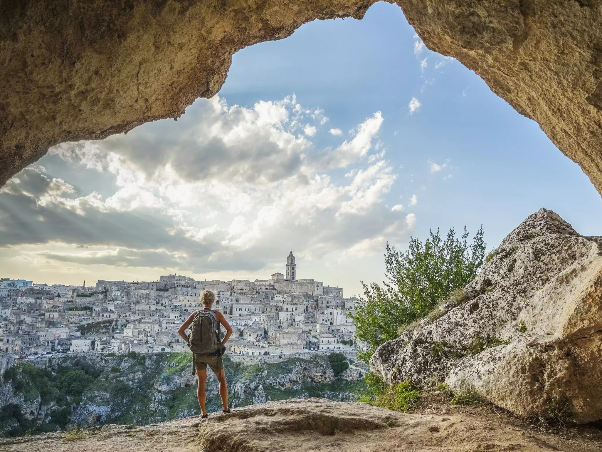 A woman wearing a backpack stands in a cave looking towards a hilltop town of white buildings on the other side of a gorge.