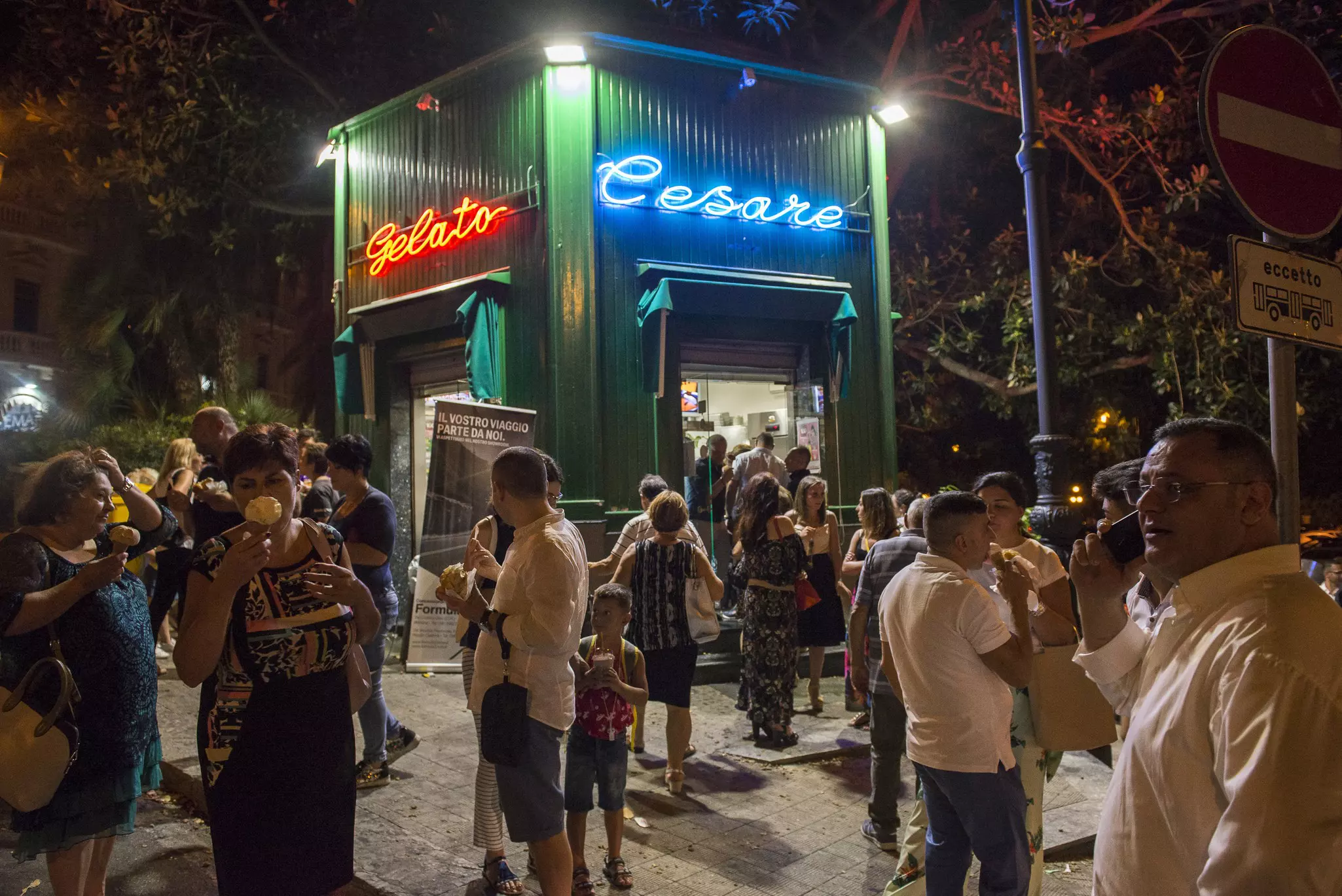 A crowd of people with ice cream cones and cups in front of an ice-cream kiosk with neon signage on a warm night
