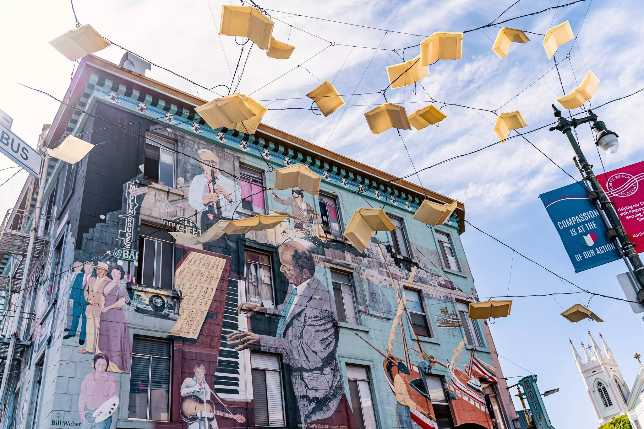 Mural of musicians, artists, and historical figures and art installation of off-white books hanging on street lines above on a partly sunny day.