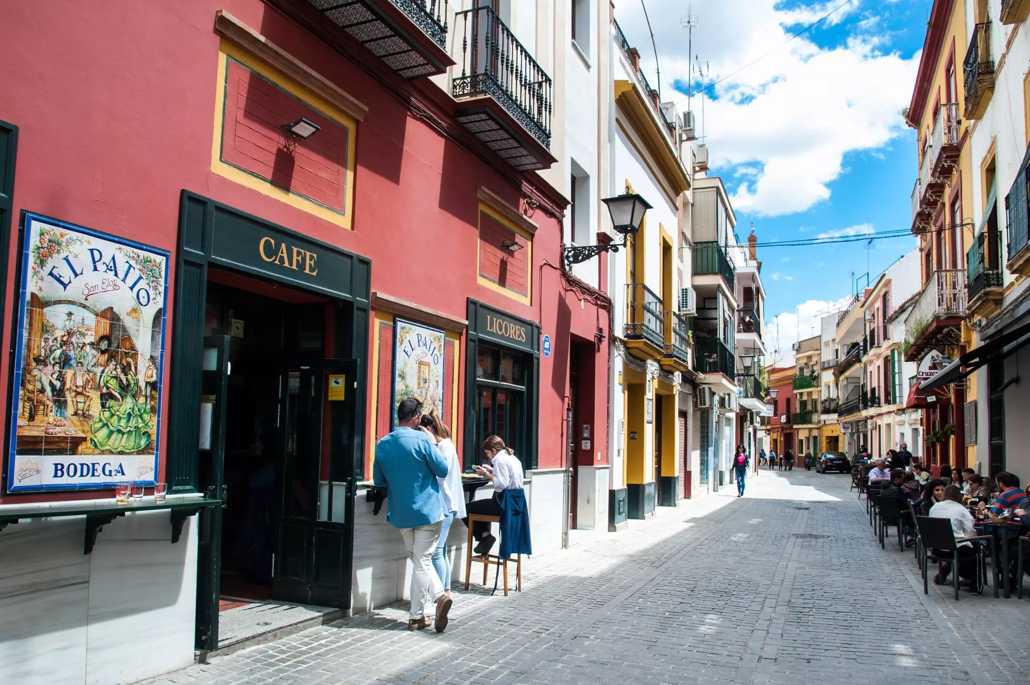 People at outdoor cafes on a narrow street with colorful low-rise buildings.