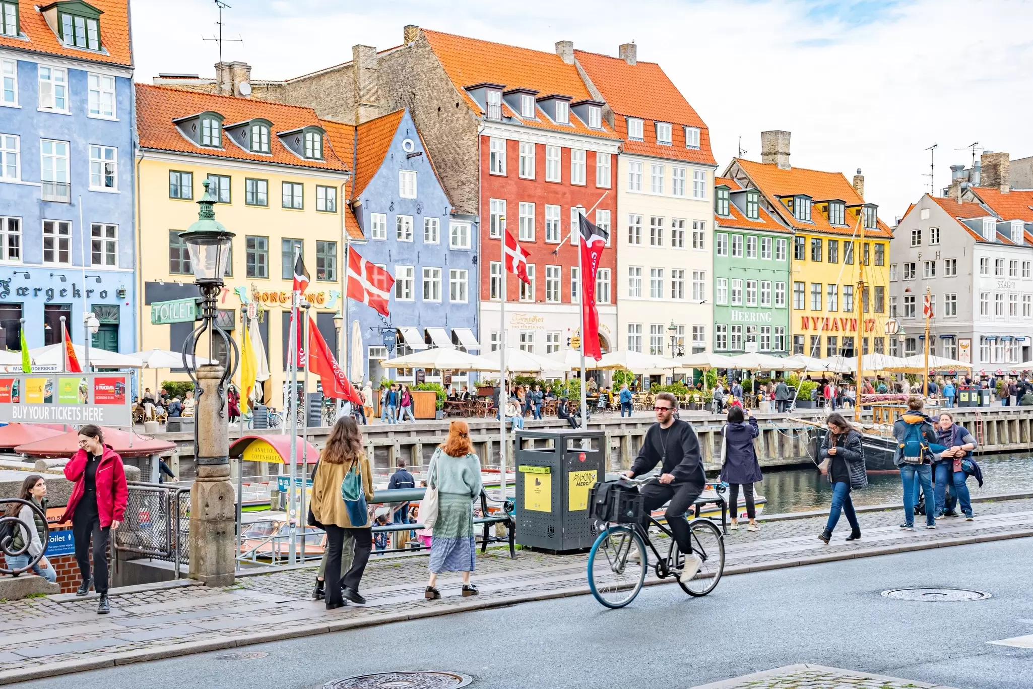 Cyclist, pedestrians and tourists in Christianshavn, Nyhavn neighborhood, Copenhagen, Denmark with traditional Danish colorful houses, boats and flags on background.