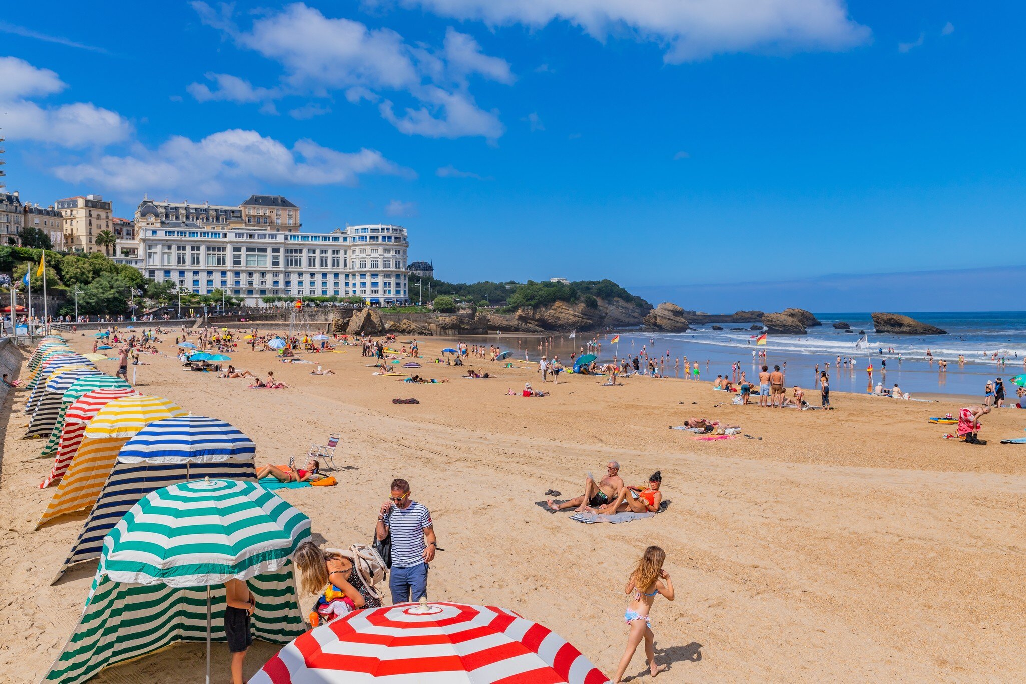 A crowded summer beach lined with brightly striped canvas tents and parasols, below a white belle-époque hotel, with rocks in the sea beyond.