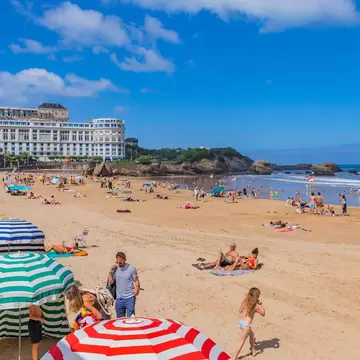 A crowded summer beach lined with brightly striped canvas tents and parasols, below a white belle-époque hotel, with rocks in the sea beyond.