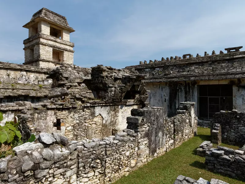 Ancient stone ruins of a low building with a tower in the background on a sunny day.