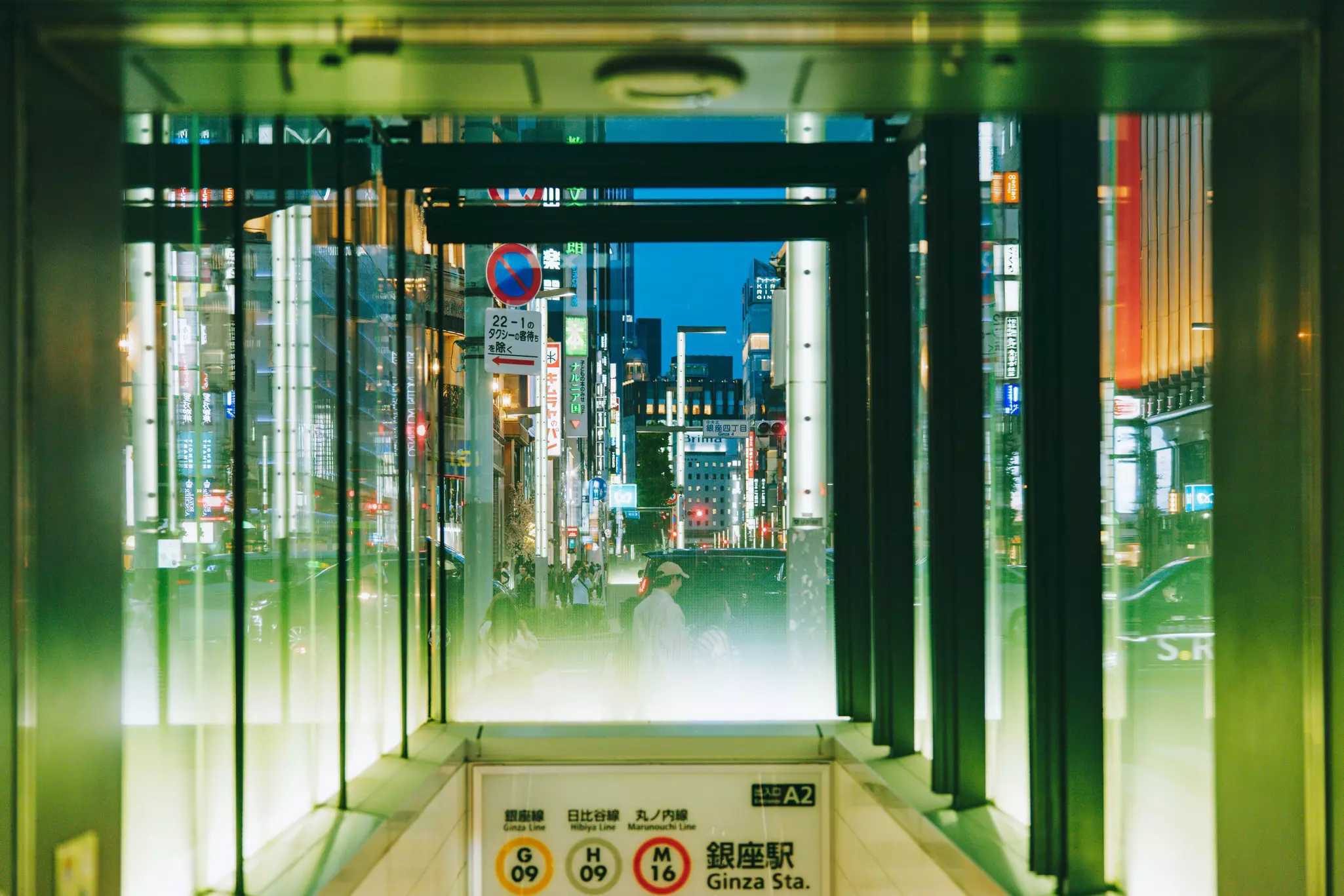 Signs lit up at night through the glass walls of a subway entrance.
