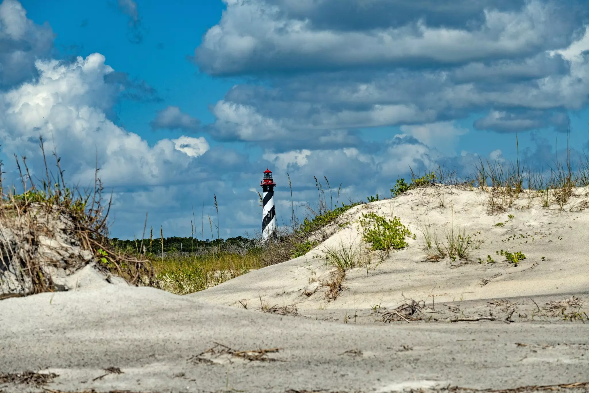 Grass grows on sloping sand dunes. A striped lighthouse is visible in the distance.
