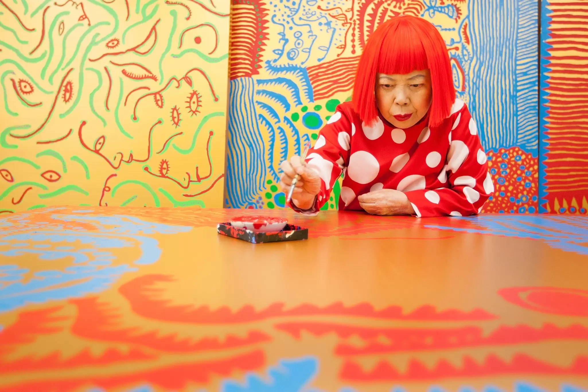 A woman, wearing a vibrant red wig in a blunt bob cut, sits in an art studio surrounded by bright colors: Yayoi Kusama.