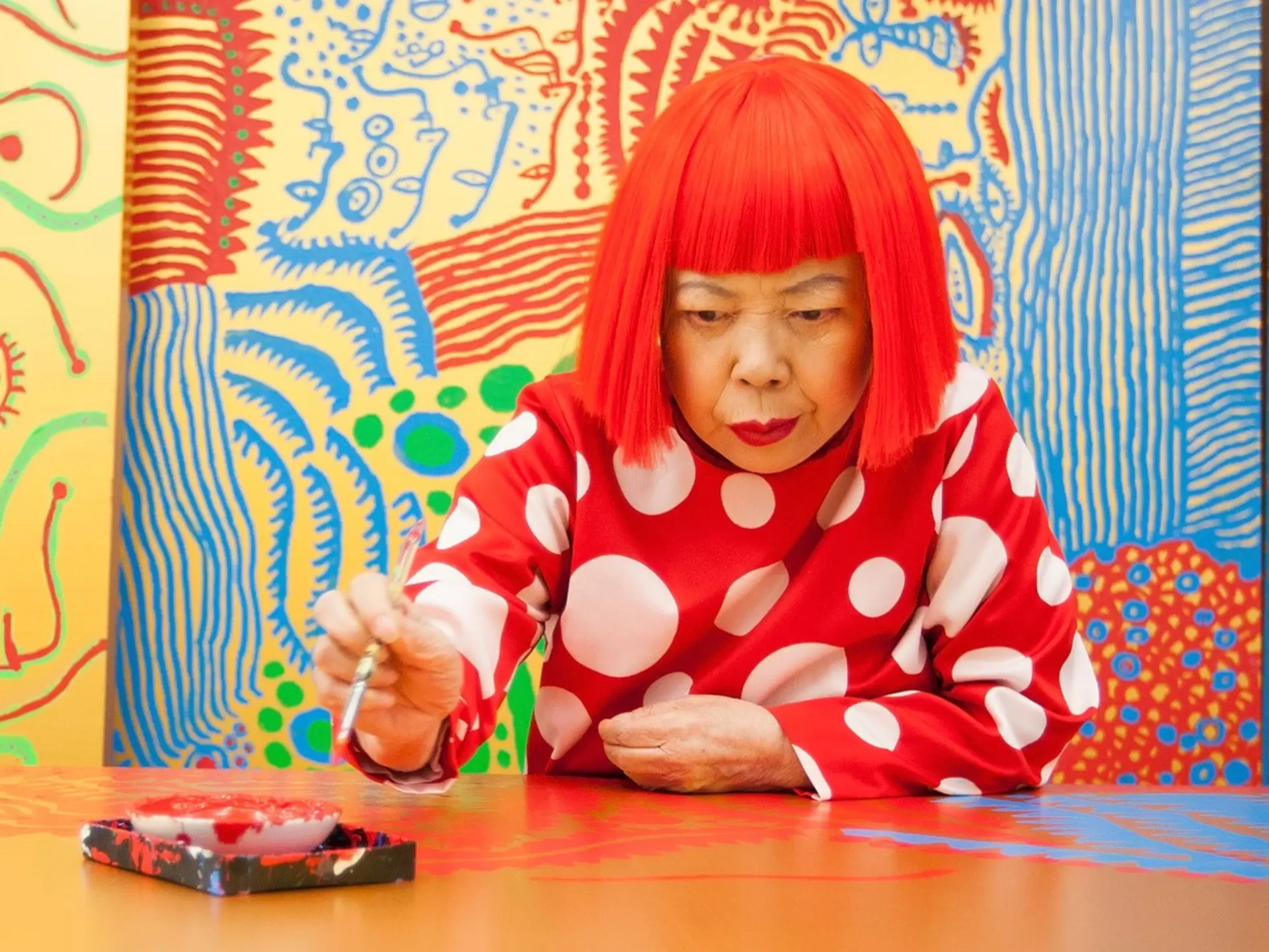 A woman, wearing a vibrant red wig in a blunt bob cut, sits in an art studio surrounded by bright colors: Yayoi Kusama.