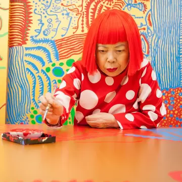A woman, wearing a vibrant red wig in a blunt bob cut, sits in an art studio surrounded by bright colors: Yayoi Kusama.