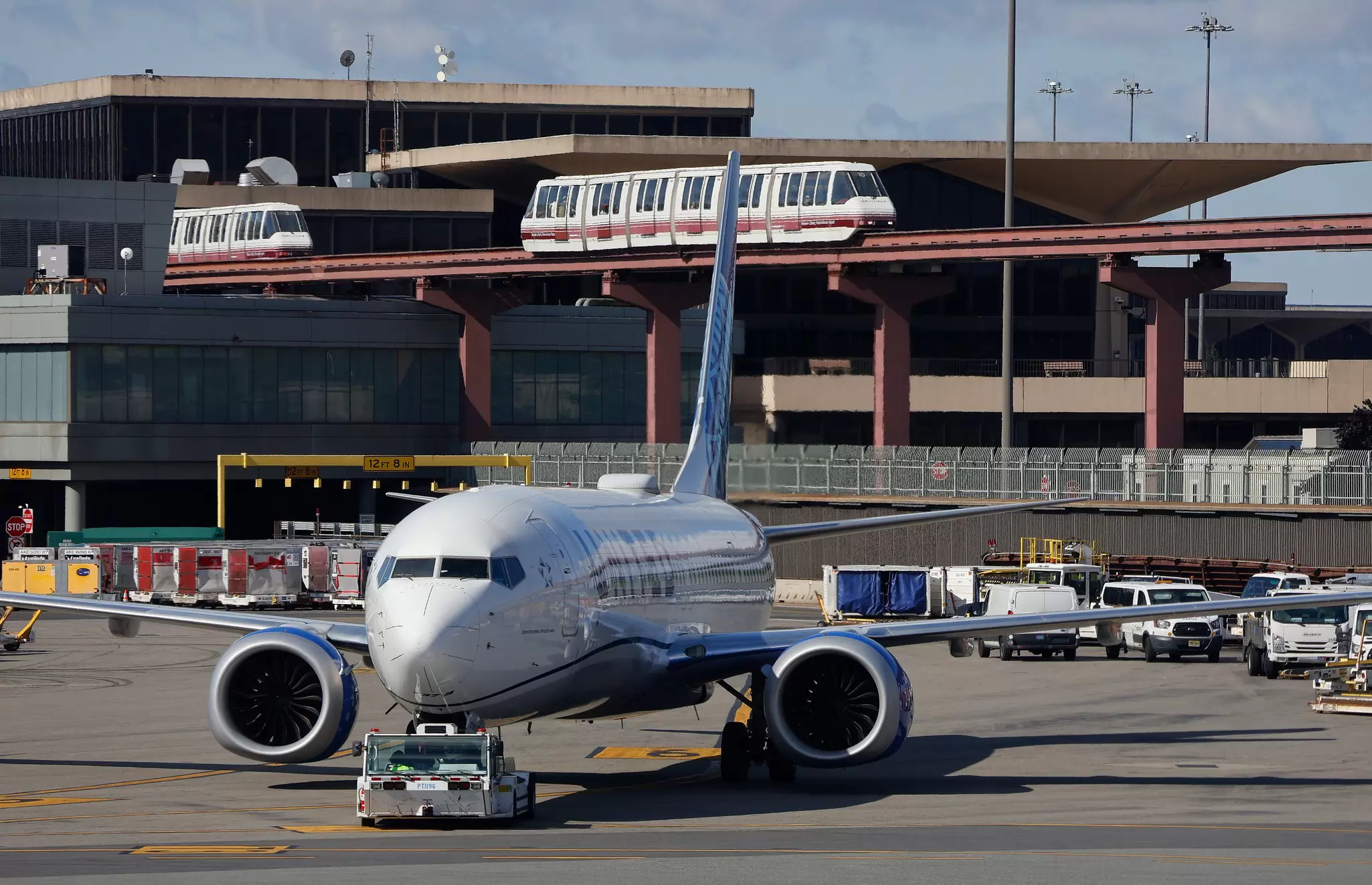 A plane is seen on the tarmac by an airport terminal. Monorails are seen passing the background.