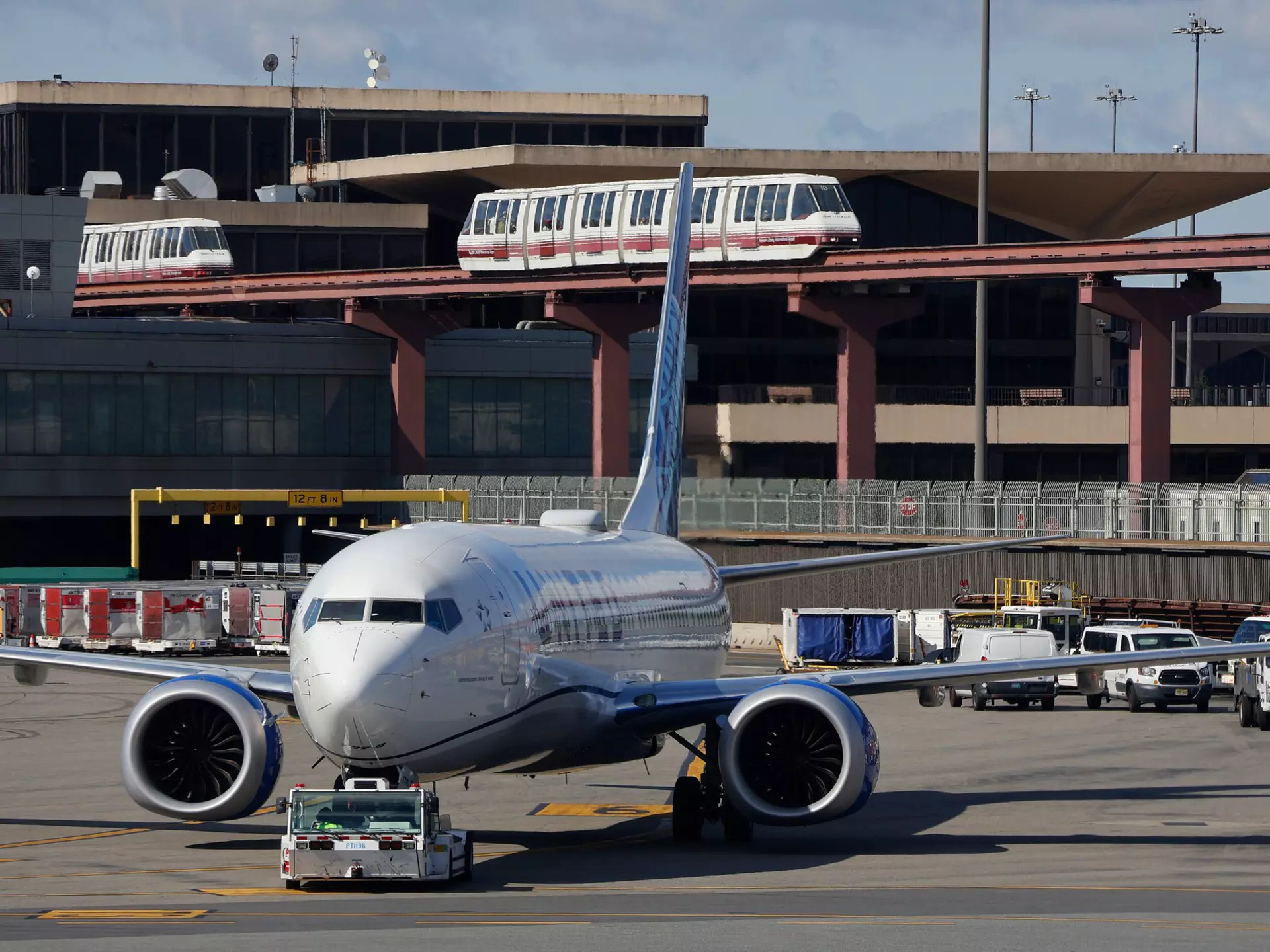 A plane is seen on the tarmac by an airport terminal. Monorails are seen passing the background.