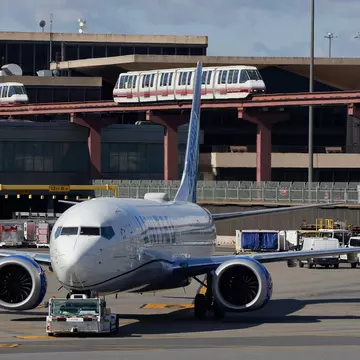 A plane is seen on the tarmac by an airport terminal. Monorails are seen passing the background.