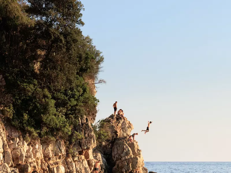 People jump off a rugged cliff into the sea as the sun sets. 
