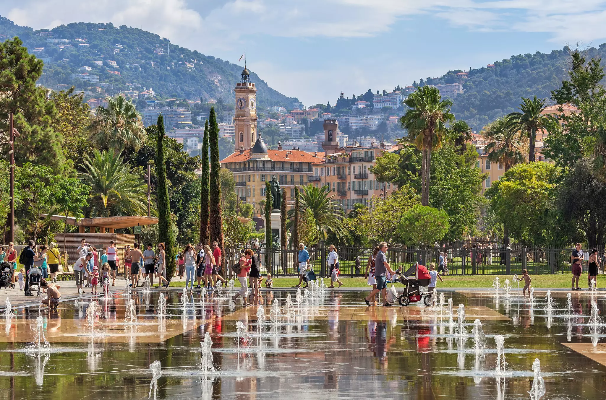 People walking and playing among fountains in a city backed by hills