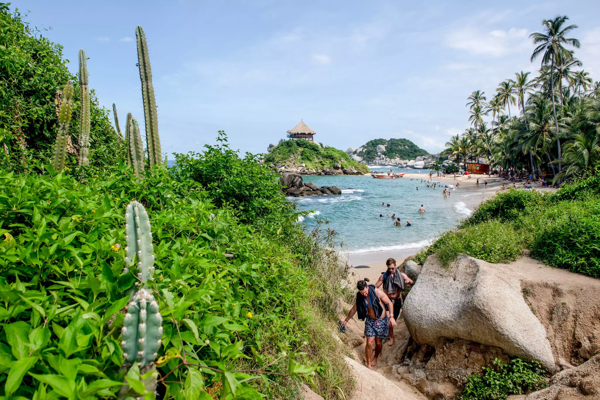 Two people clamber over boulders near a sandy path leading away from a tropical beach on a sunny day.