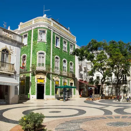 Looking over a sunlit square, paved with stones in a circular geometric pattern; the centre of attention is a handsome green tiled house, alongside white buildings and trees.