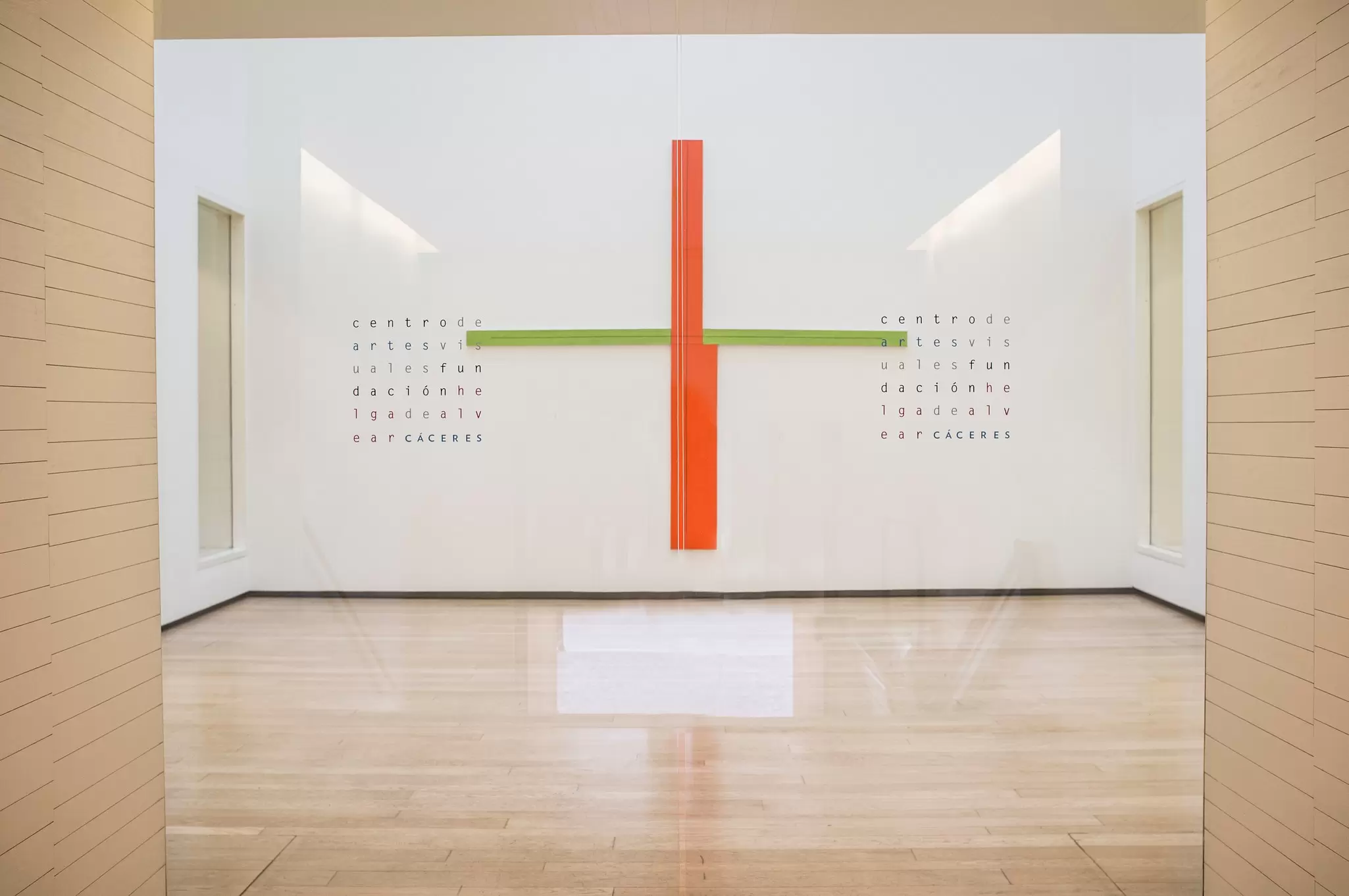 A bright white gallery with pale wood floors and a large red and green cross artwork on the end wall.
