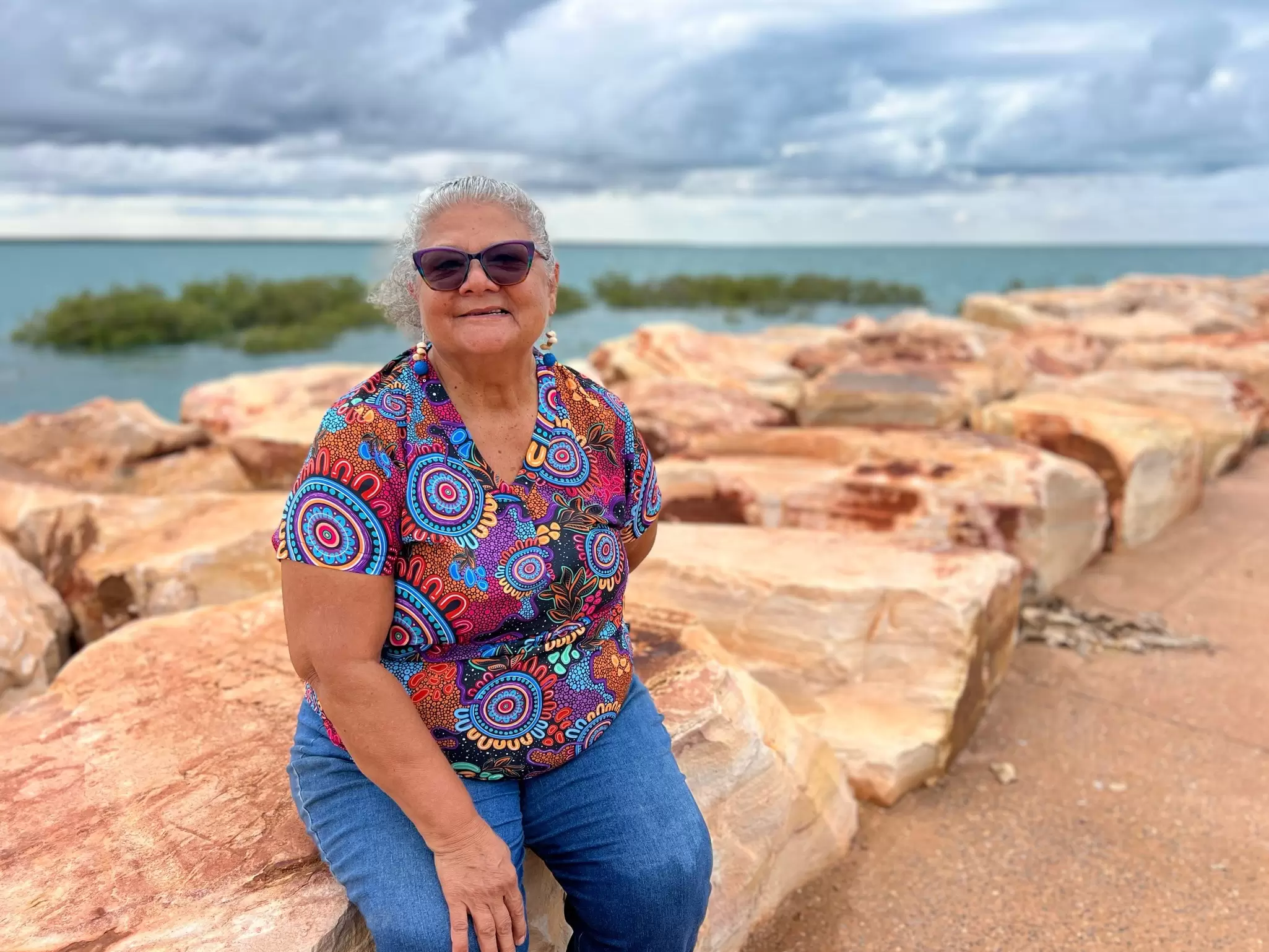 An Aboriginal woman in a colorful Indigenous motif shirt sits on a wharf.