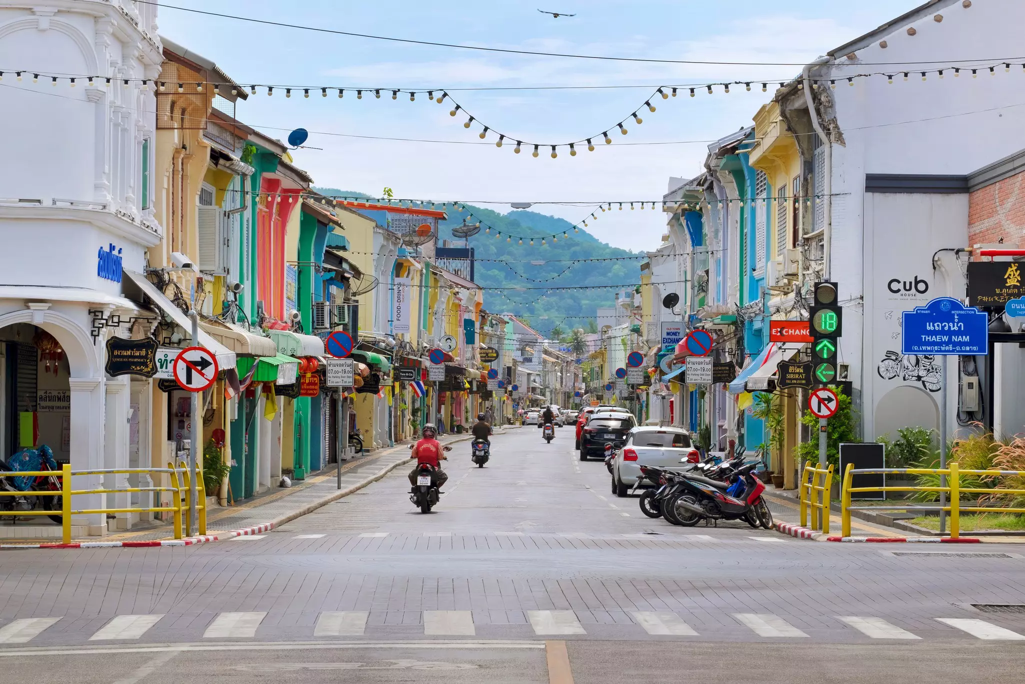 Motorcyclists ride down a small street of pastel-colored buildings and shops.