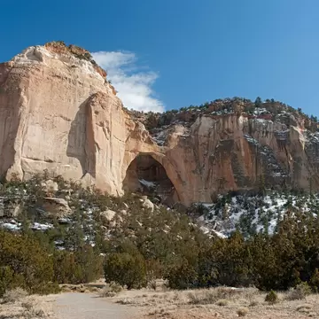 Large natural arch in pale sandstone cliffs with patches of snow, above junipers and a dirt trail in El Malpais, New Mexico