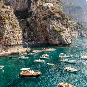 Motorboats and wooden launches on turquoise water below sheer limestone cliffs, a small beach with sunbeds at the back