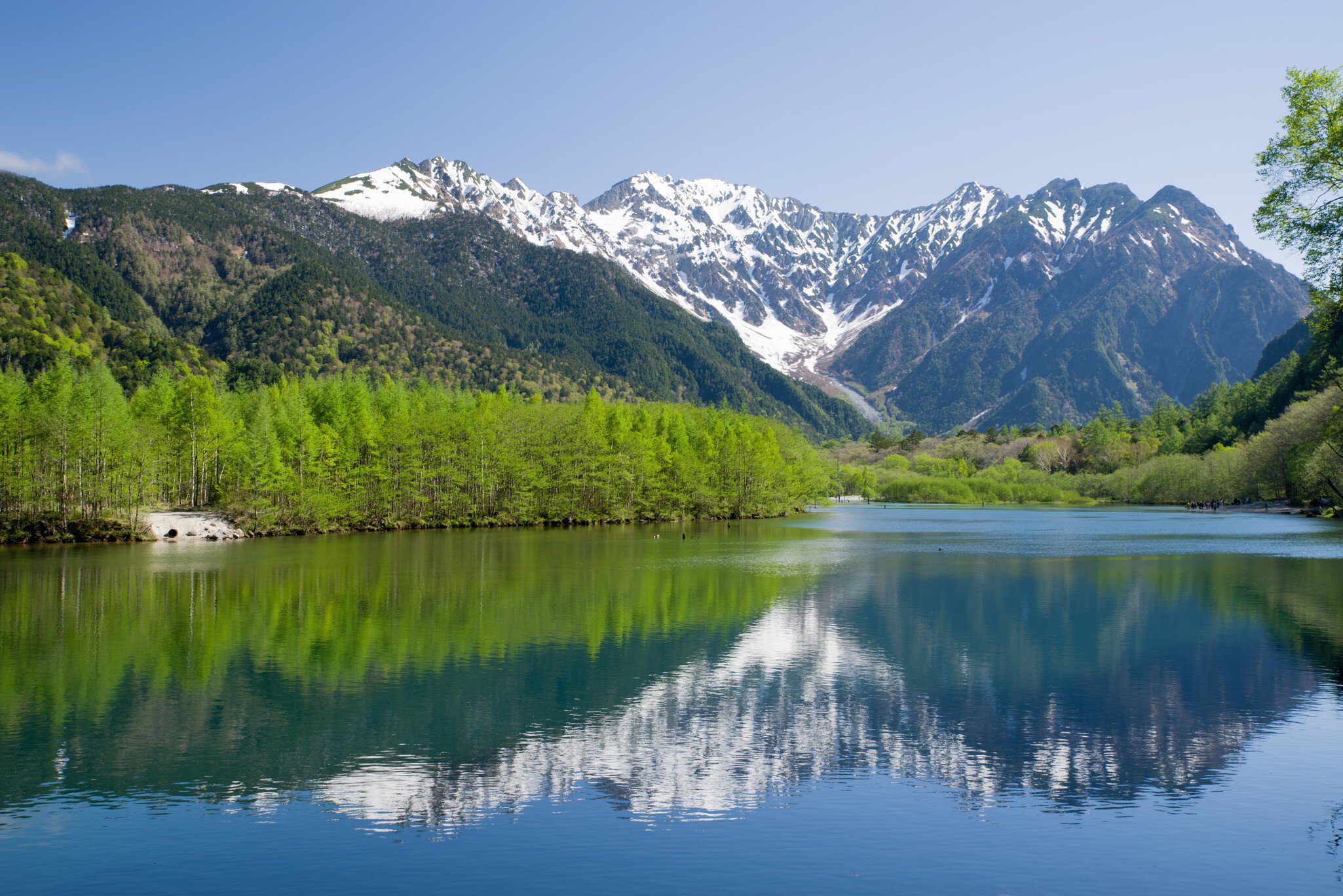 Rocky, snow-covered slopes are reflected in a blue pond with green trees on the shore.