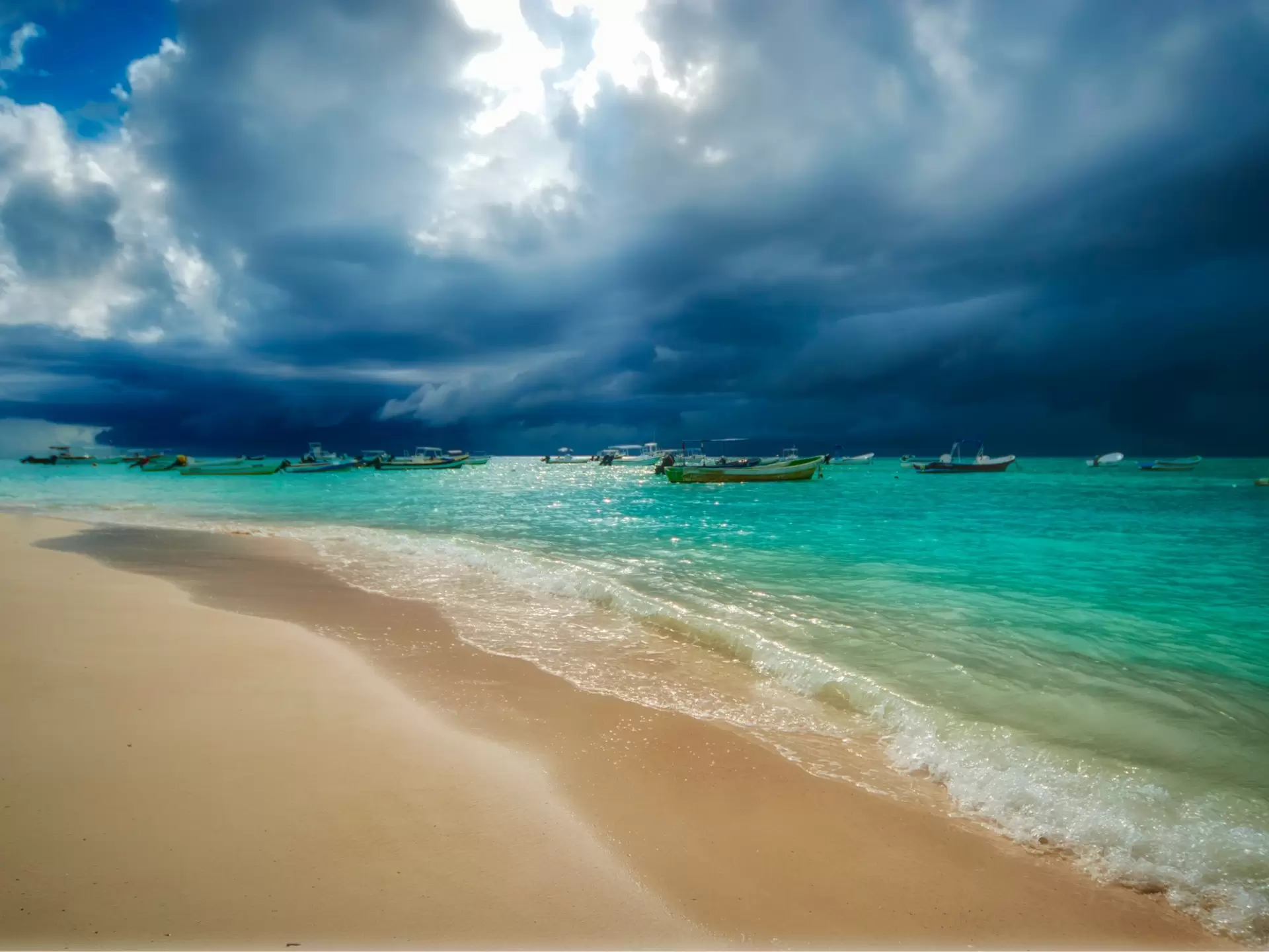 Storm clouds gather out at sea over a beach, with small boats moored in the water