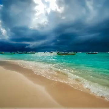 Storm clouds gather out at sea over a beach, with small boats moored in the water