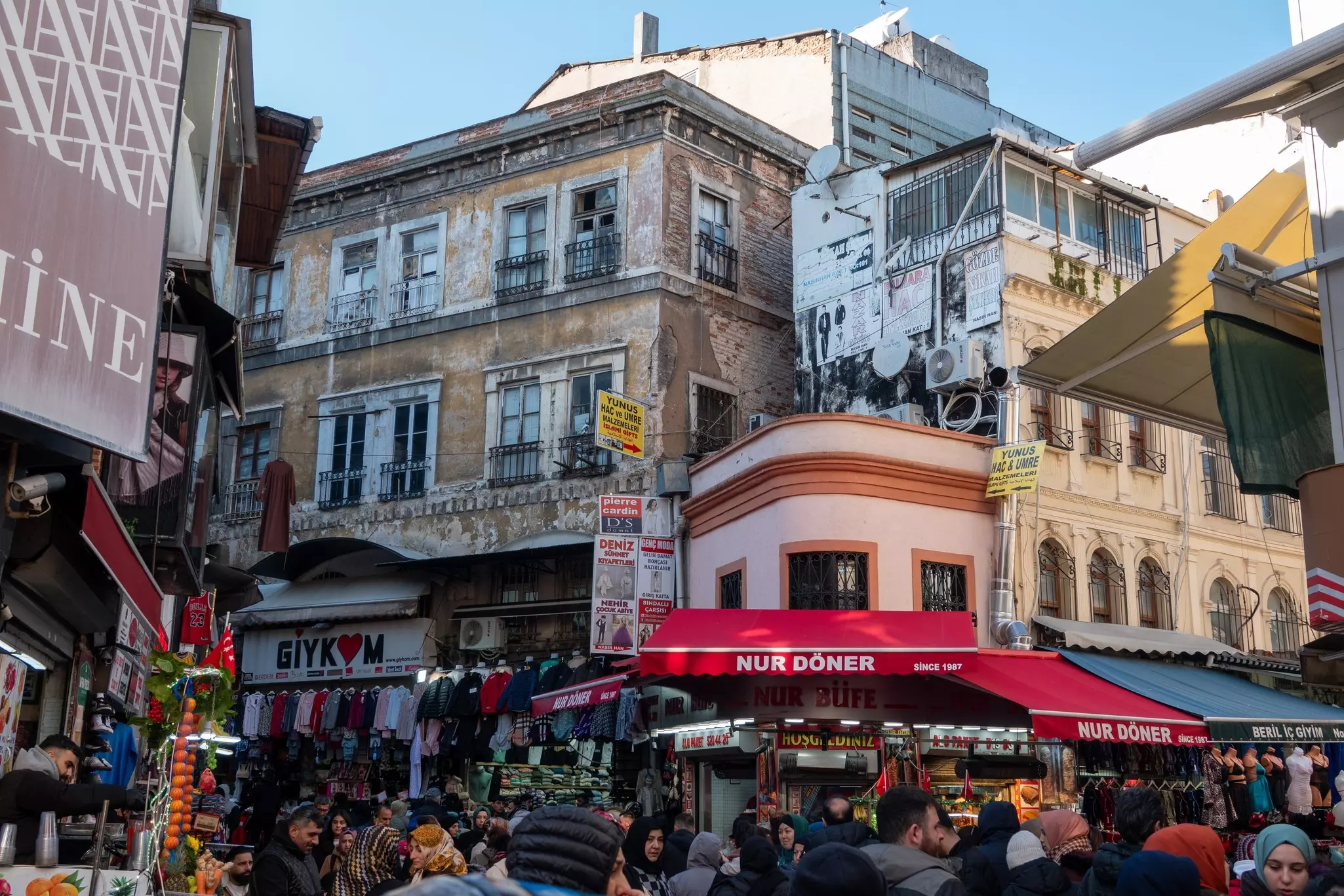 People shopping on a crowded street surrounded by old stone buildings on a sunny day.