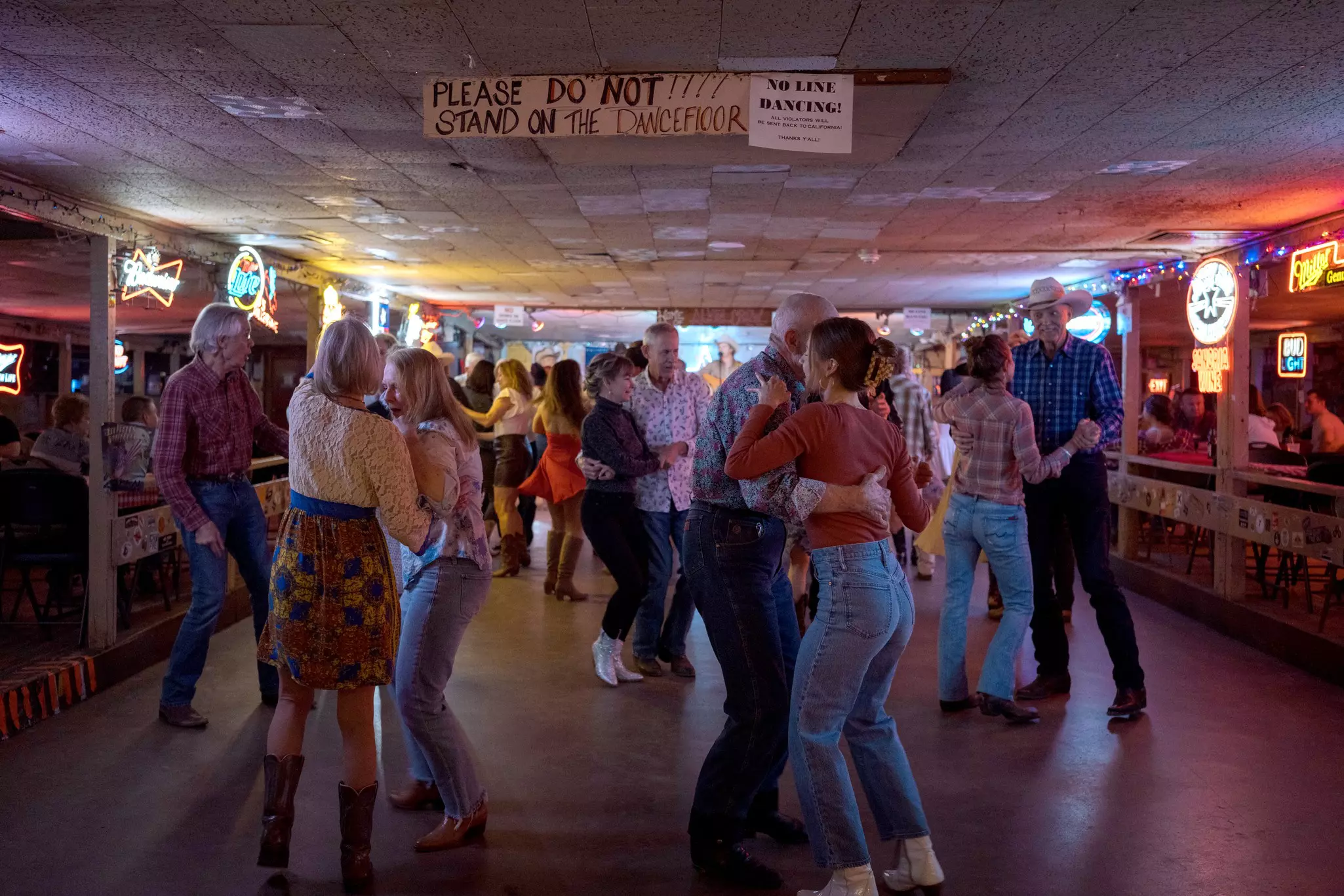 Couples dance on the dance floor of a bar with neon signs.
