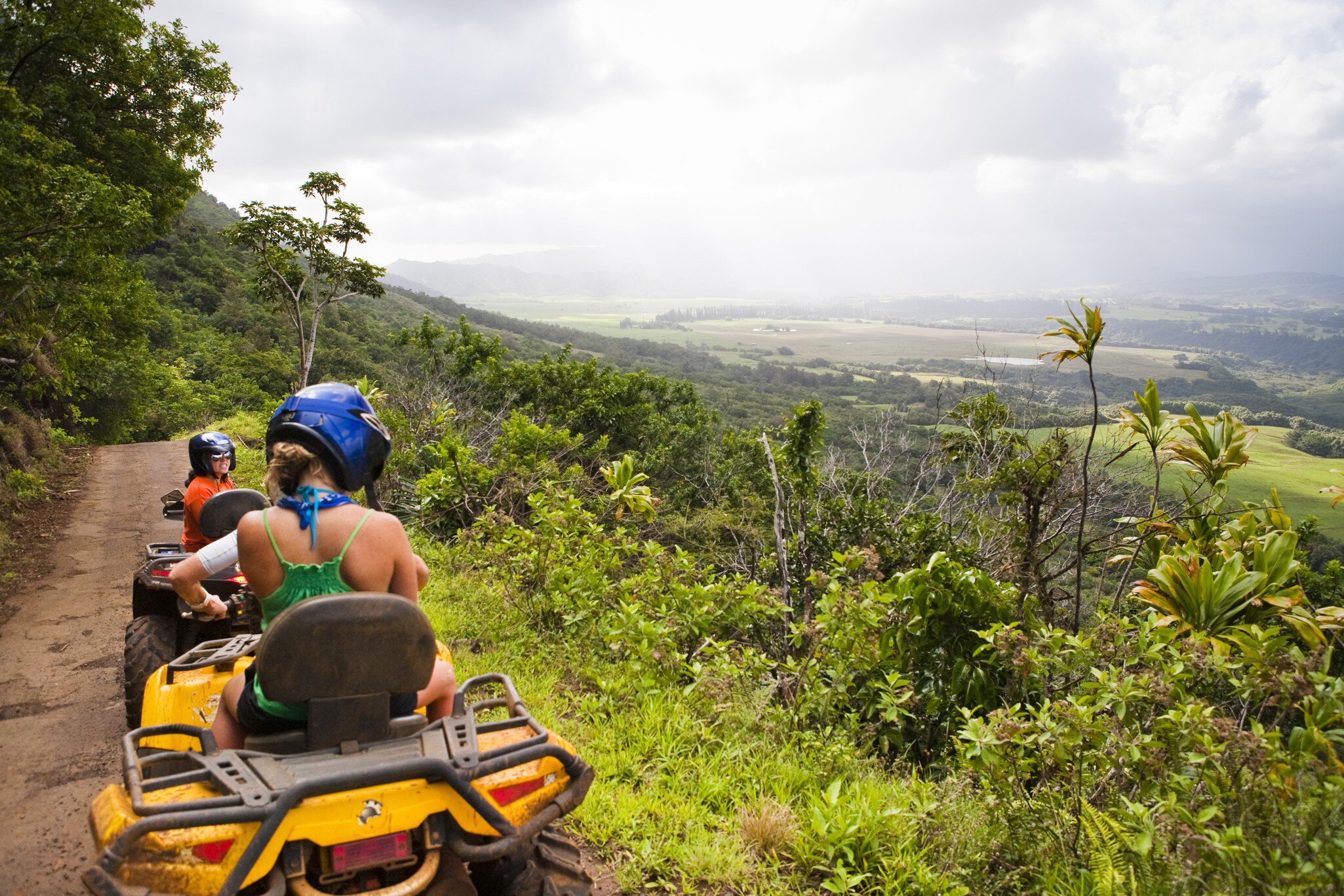Two people in helmets ride ATVs along a muddy ridge trail overlooking a broad green valley and misty mountains