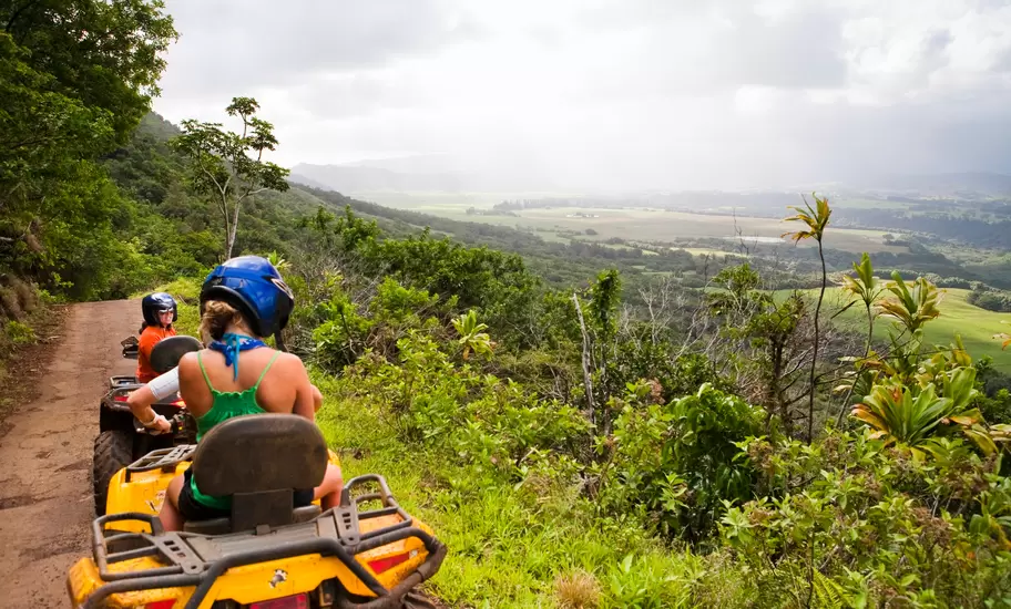 Two people in helmets ride ATVs along a muddy ridge trail overlooking a broad green valley and misty mountains