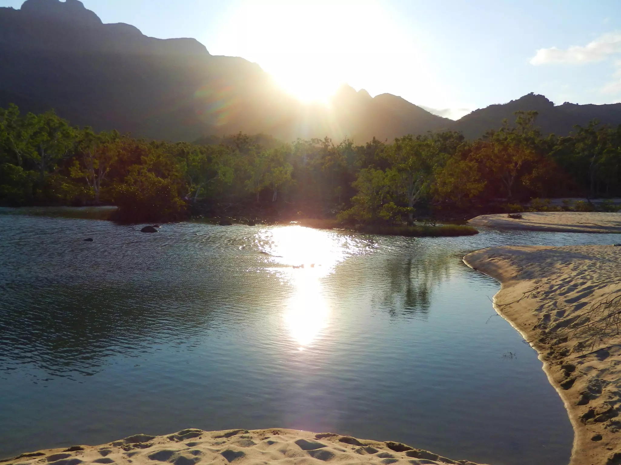 The sun peeps over a mountain top creating glare off a lake