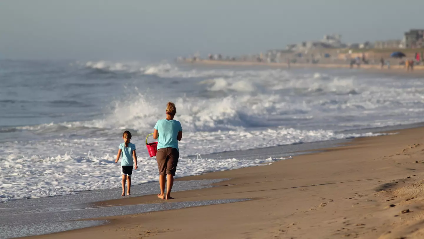 A mother and her young daughter walk along a beach in the morning. The waves are high and the mother is carrying a red bucket
