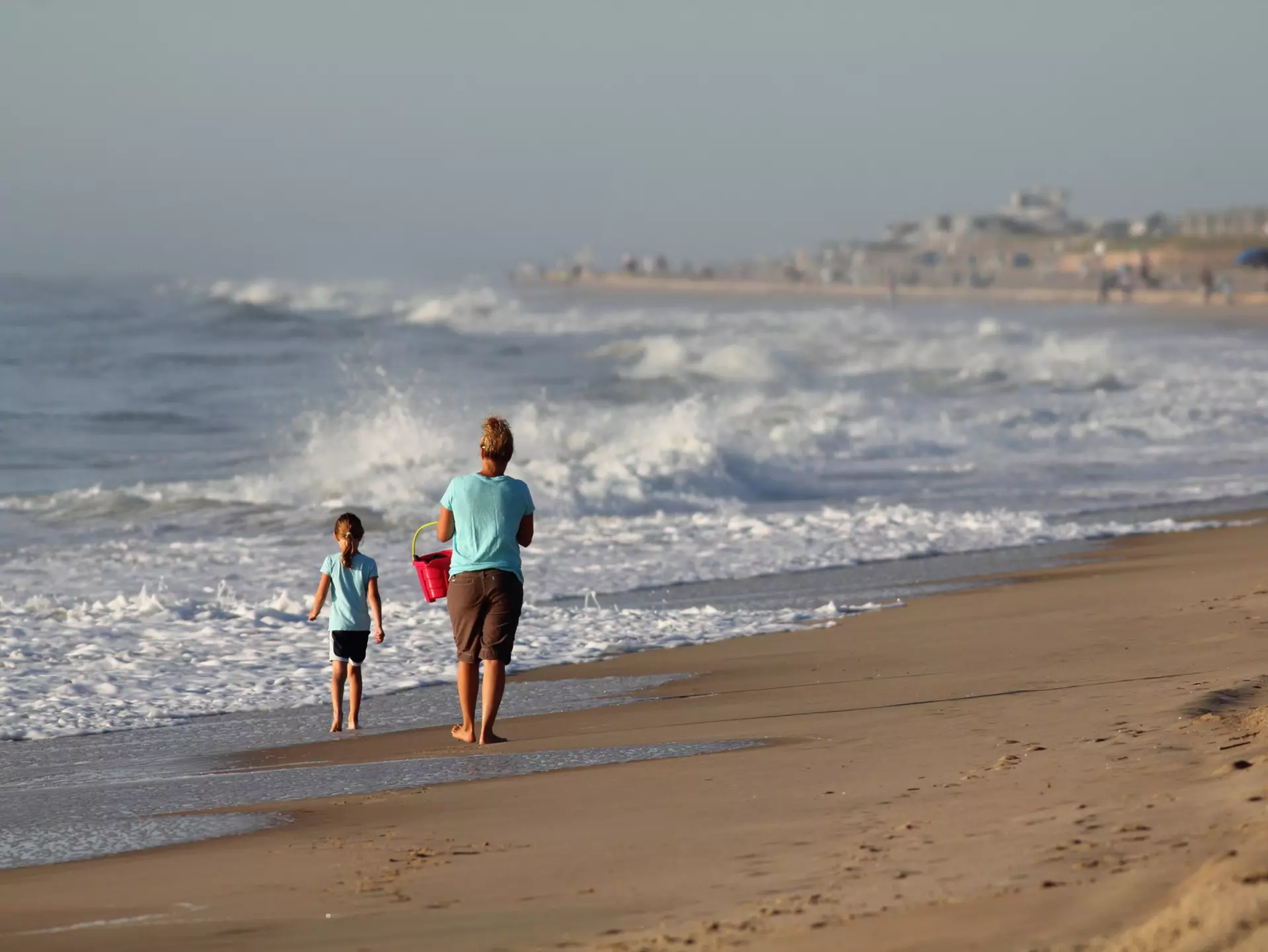 A mother and her young daughter walk along a beach in the morning. The waves are high and the mother is carrying a red bucket