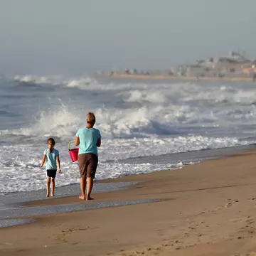 A mother and her young daughter walk along a beach in the morning. The waves are high and the mother is carrying a red bucket